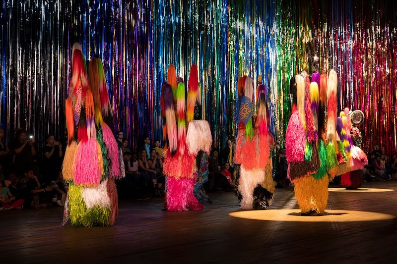 Nick Cave's color Soundsuit performers are enshrouded in his neon raffia, staffs of hair or fur. They stand in front of the mylar streamers.
